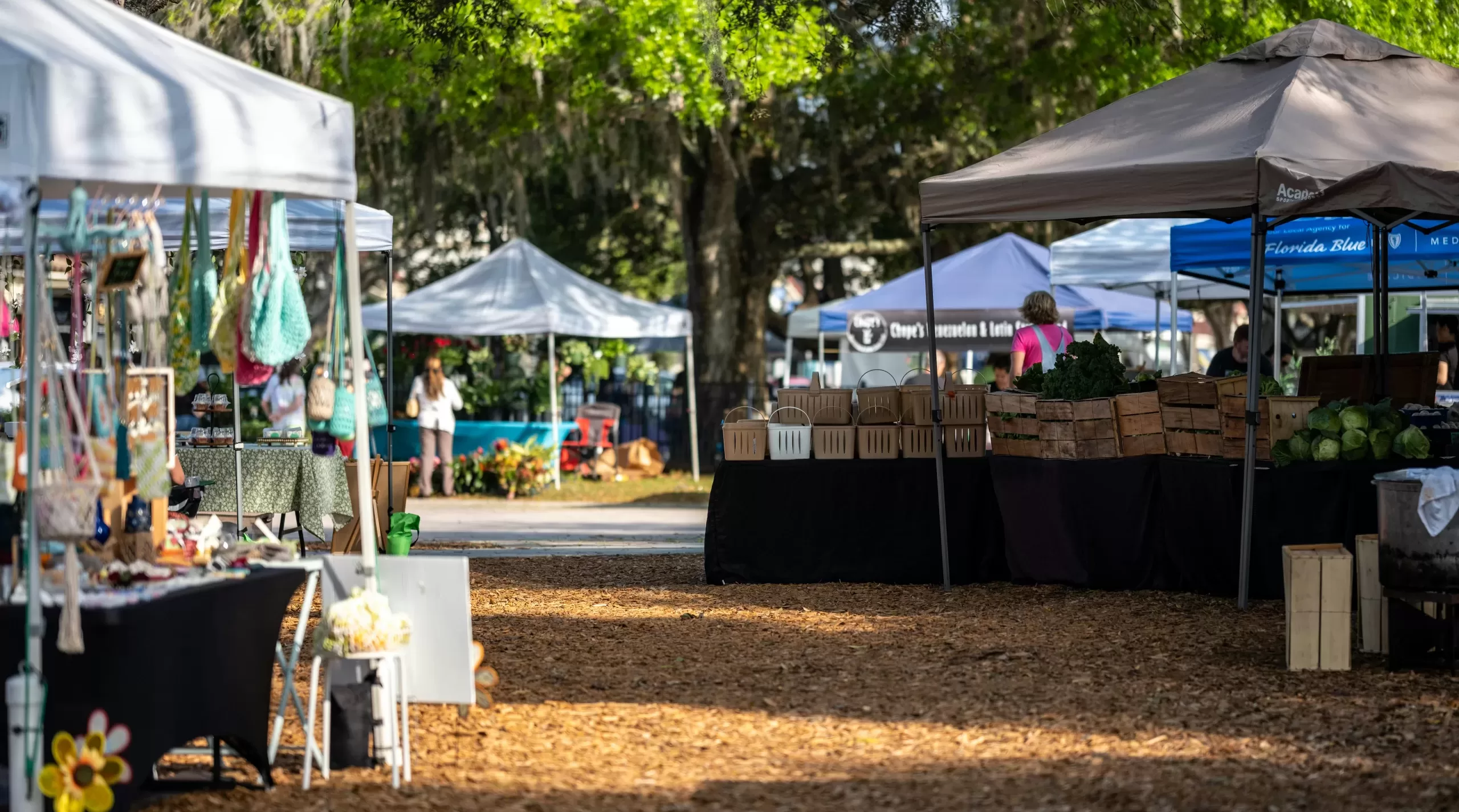 A lively outdoor market scene with vendor stalls and shoppers under trees.